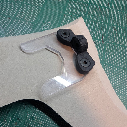 A close-up of a clear plastic visor or lens protector with two black rubberized mounting pieces, placed on a beige sheet over a green cutting mat.