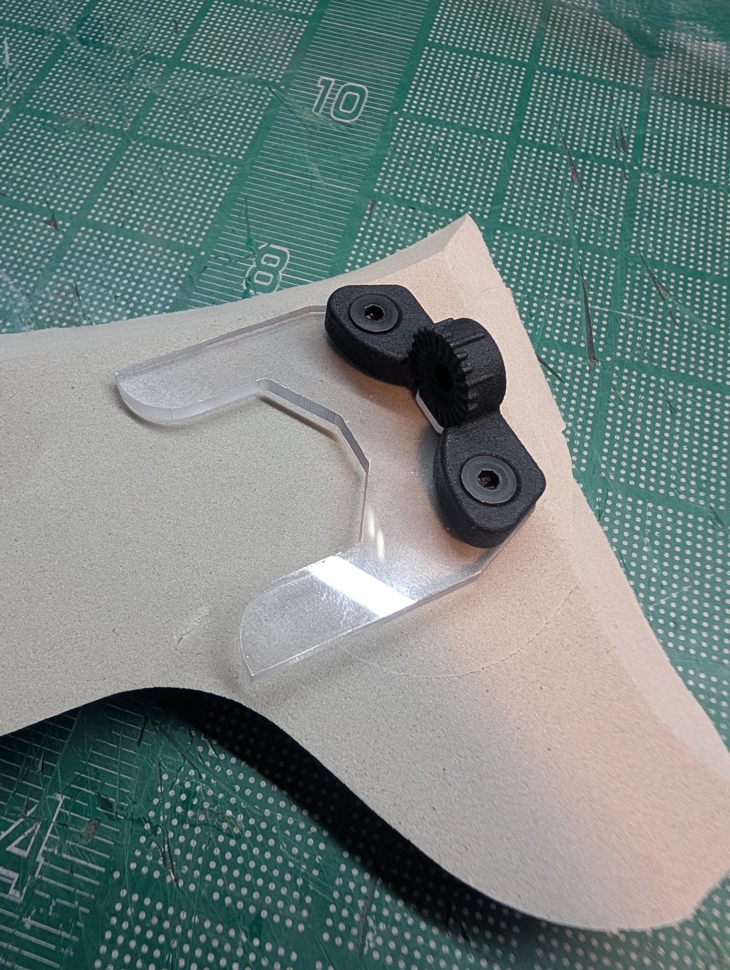 A close-up of a clear plastic visor or lens protector with two black rubberized mounting pieces, placed on a beige sheet over a green cutting mat.