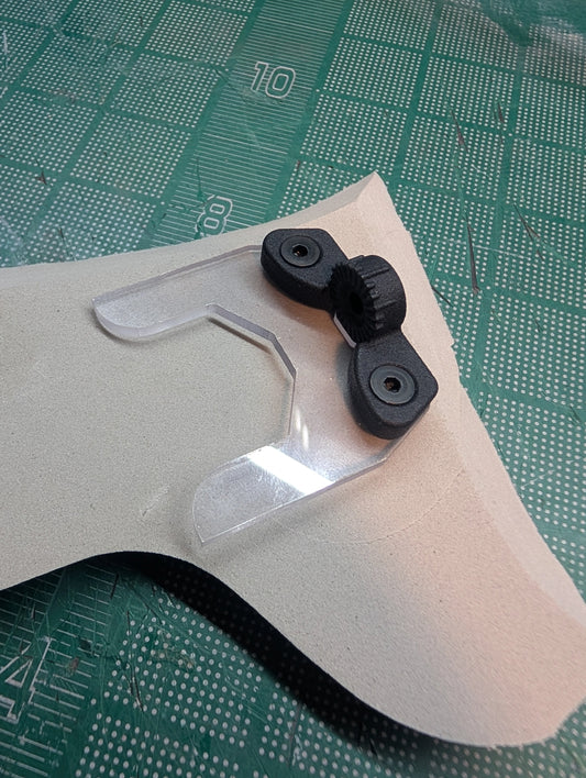 A close-up of a clear plastic visor or lens protector with two black rubberized mounting pieces, placed on a beige sheet over a green cutting mat.